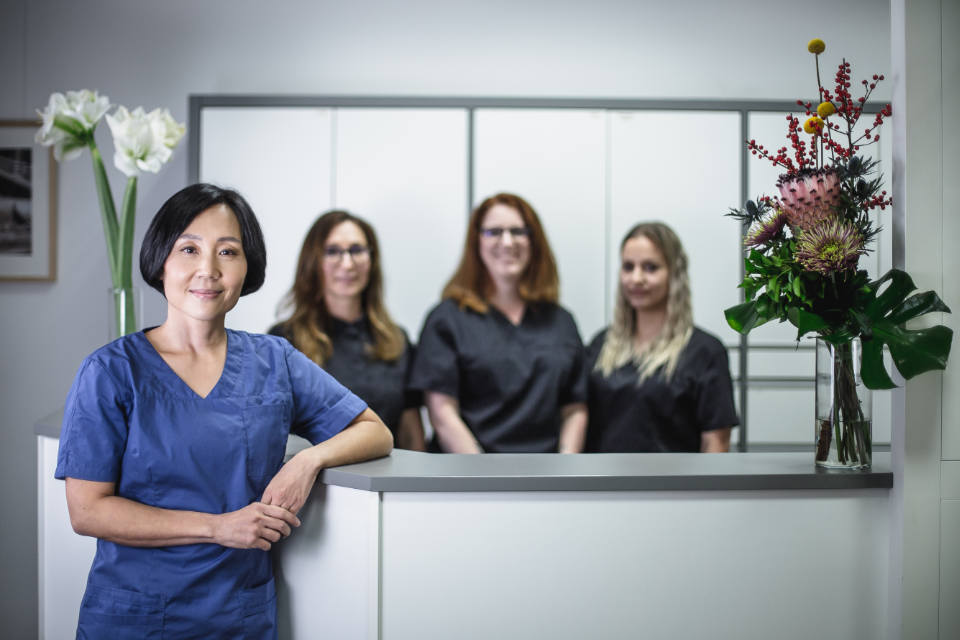 Dr. Sonja Tai in blauer Arbeitskleidung im modernen Praxisempfang, drei Teammitglieder im Hintergrund, dekorative Blumenvasen.